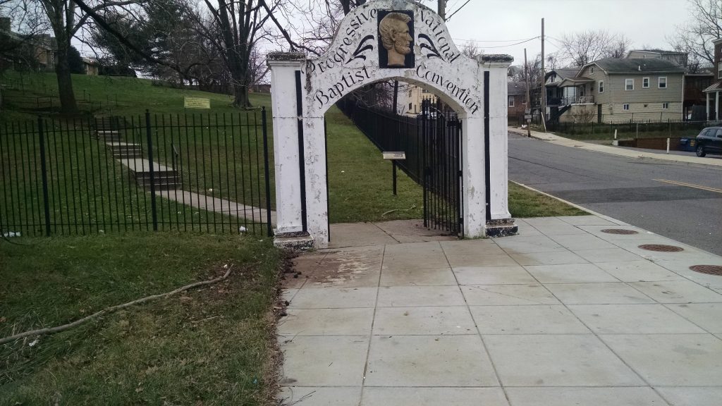 Front white gate of a fence with peeling white paint. THere is an gold image of Lincoln on top of the arch and it reads "Progressive National Baptist Convention" in black cursive font. 