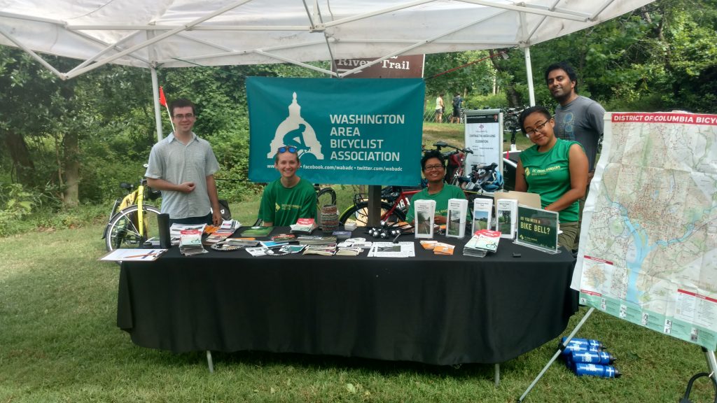 5 people behind a table with lots of outreach material. There is a WABA logo banner behind them, they are under a tent outside on the grass and three of the people are wearing green Trail Ranger shirts. There are 3 men and 2 women. 