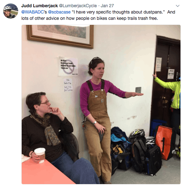 Screen caption of a tweet reading "@WABA's @sobacase "I have very specific thoughts about dustpans." And lots of other advice on how people on bikes can keep trails trash free." There is a photo of a woman in brown workpants overalls talking and leaning up against a wall. Another woman on the left is thinking, a pile of bike gear is on the right with the arm of a person is high visibility jacket on the far right of the frame. 