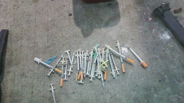 many hypodermic needles in a pile on a concrete floor. Clearly from a cleanup. 