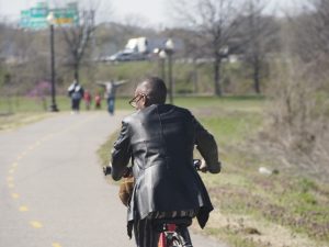 Black guy riding a Capital Bikeshare bike and a fancy leather jacket bikes away from the camera in Anacostia park. You can see a few other trail users in the background