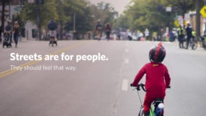 A child in a superhero costume rides a bike down car free Georgia Avenue during DC's first open streets event. Text reads "Streets are for people. They should feel that way."
