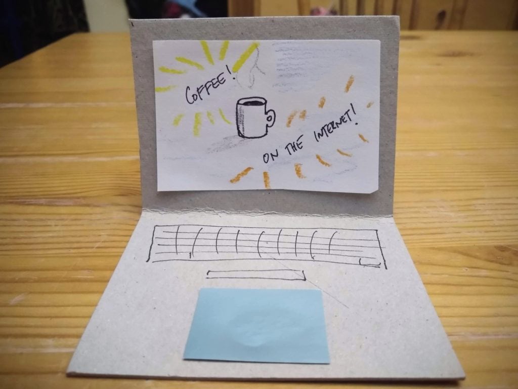 A fake cardboard laptop sits on a table, the screen says Coffee! On the Internet! with a drawing of a mug