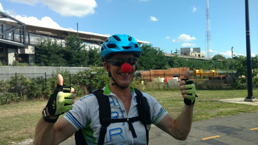 A white guy in spandex wears a red clown nose and a blue helmet and is giving two thumbs up to the camera with a big smile