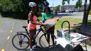 A Black woman trail ranger stands next to a trail holding a map talking to a Black woman on a road bike. It is sunny and the middle of summer.