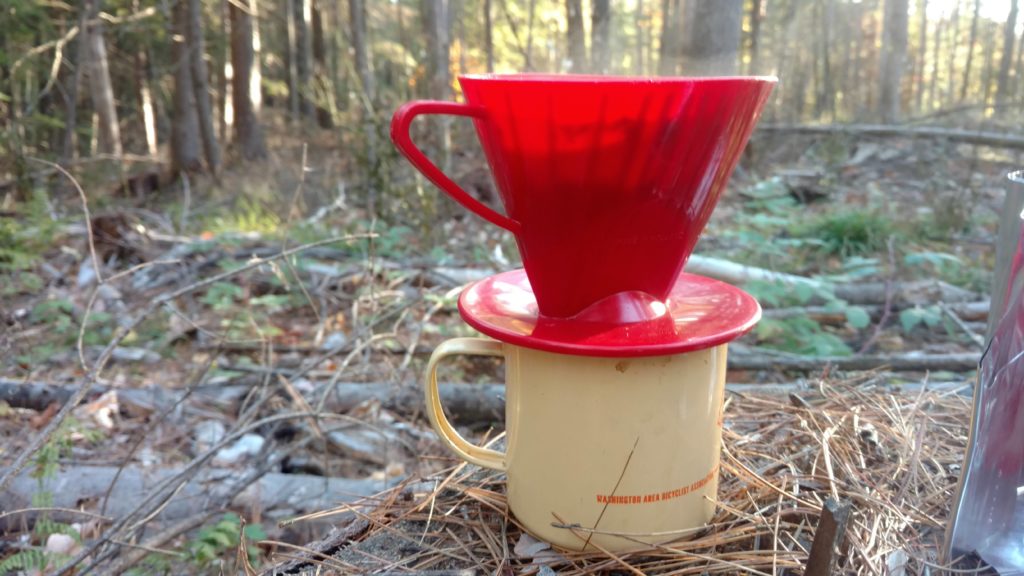 Chipped WABA mug sits on the forest floor with a coffee filter above and steam coming out