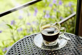 Espresso in a clear glass cup sits on a table, there are lavender flowers behind. It is and looks like a stock photo