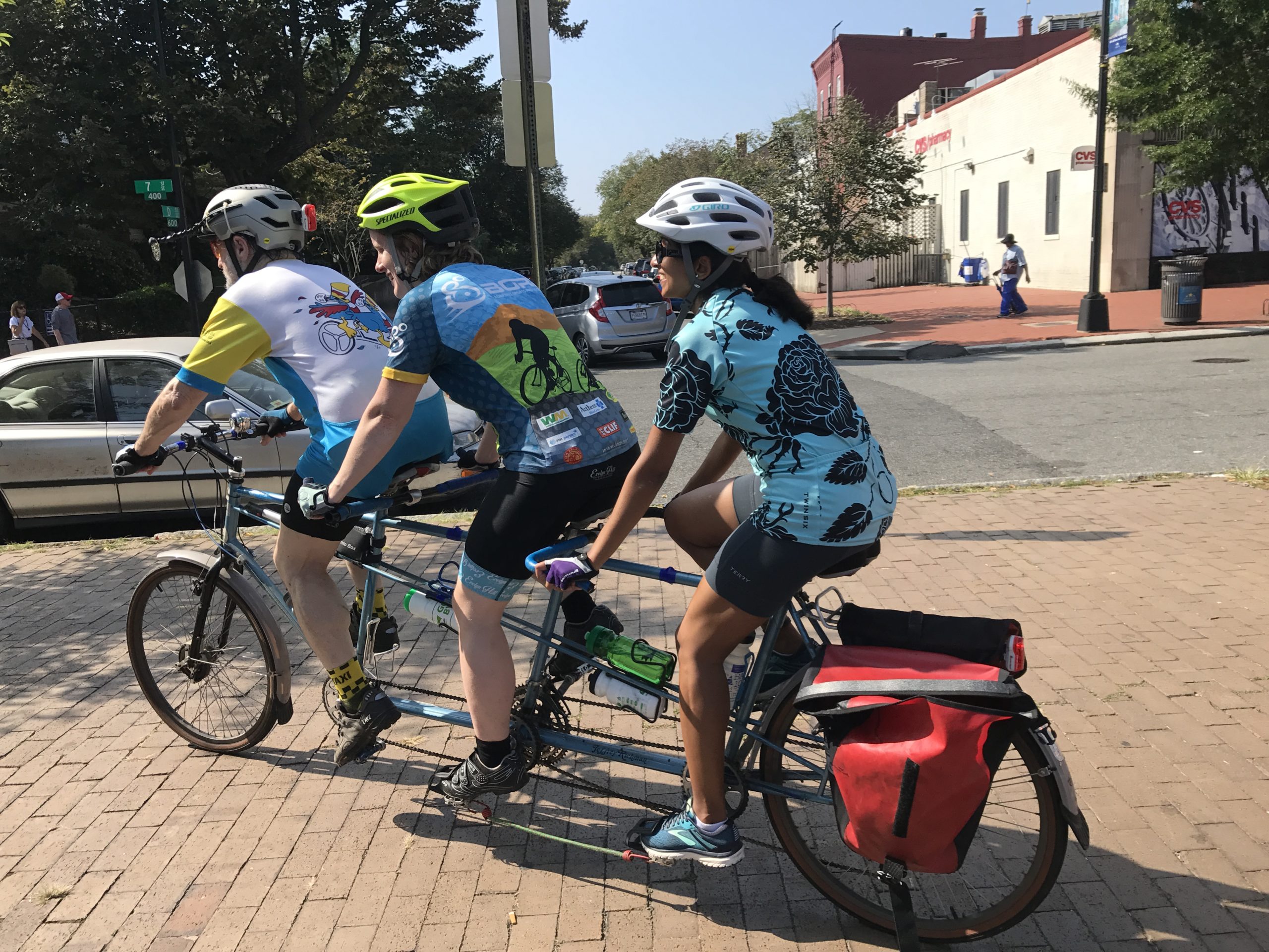 Three people are riding in a line on a three person bicycle away from the camera so you can only see their backs. The captain is wearing a white jersy, and the two people behind are in blue jerseys. Everyone is smiling, the sky is blue and they are about to go for a ride. Photo credit: MWABA