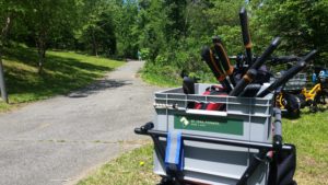 A grey boxes sits on a bicycle and is full of pruner handles. The trail is in the background, and the trees are very green. In the background you can kinda see a Trail Ranger standing in a green shirt
