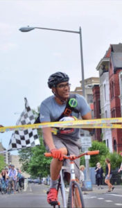 David Confer is riding a bright pink handlebar fixie as he crosses the finish line tape (its actually caution tape) on a road near Union Market area. He is smiling and is an older photo before he got cancer.